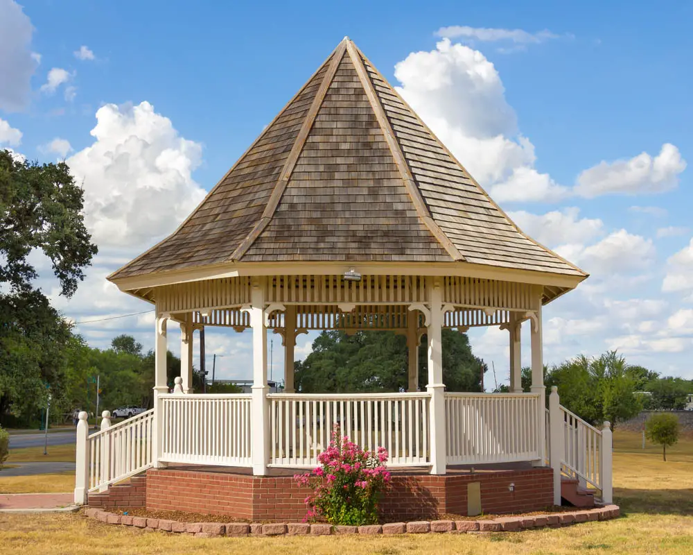 The high-peaked roof on this bespoke gazebo gives it an instant sense of charm and style, standing out among the sparse trees on this flat landscape. Built over a charming red brick foundation, wrapped in a slim garden lined with more red bricks, we see the white fencing and columns rise toward the sharply angled roof. It's a more ostentatious gazebo, perfect for luxurious landscapes.