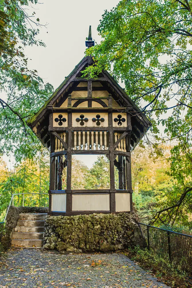 While there's a large variety of gazebo styles out there, we don't often see Bavarian inspired models. This unique structure bears all the hallmarks of its inspiration, with a two-tone design of off-white panels and dark brown beams. Additionally, the roof structure is intricately crafted, while the foundation is set into a massive stone mound.