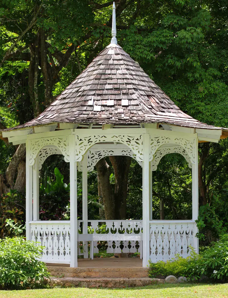 We love the ornate carving on the fence posts wrapping this gazebo, paired with the paper-cutout look of the arches between support pillars above. The pristine white paint contrasts with the earthen foundation, while the rustic wood roof adds a sense of timeless charm. It's topped with a unique spear design that helps it stand out.